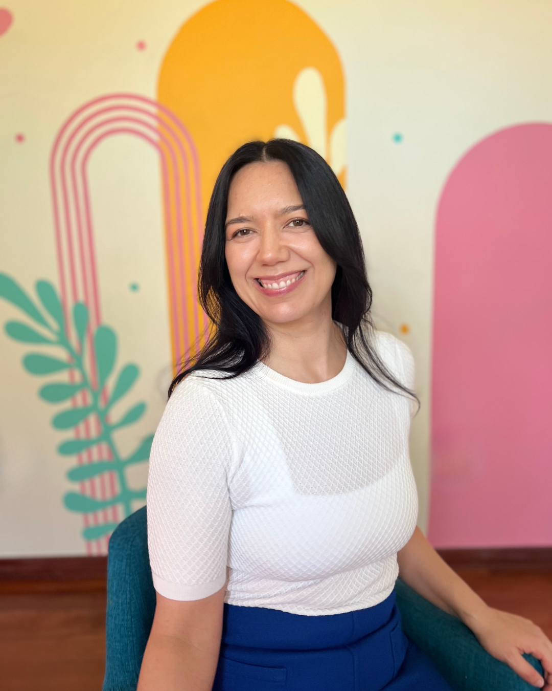 Hypnotherapy Miami - Woman with long black hair smiling while sitting on a blue chair in front of a colorful abstract background.